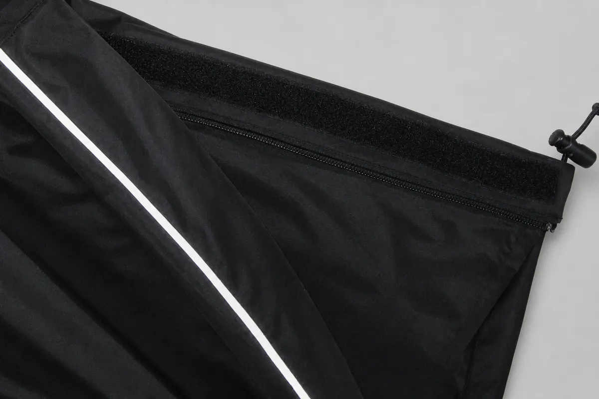 Black nylon rain-pants opening with zipper hook-and-loop adjuster drawcord and reflective piping